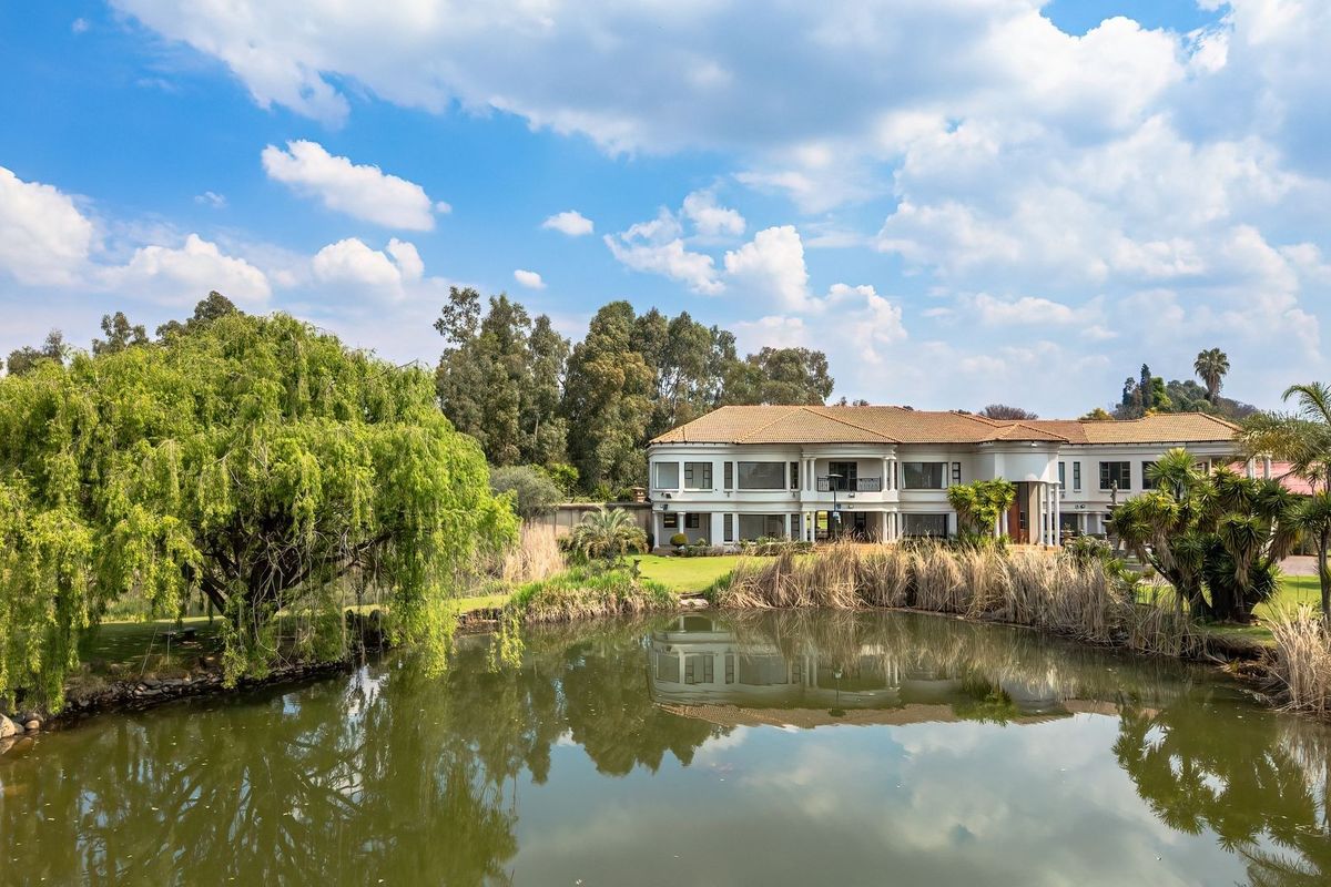 A scenic rear view of a large multi-story residence featuring a tiled roof, balcony, and columns, overlooking a tranquil pond and landscaped garden.