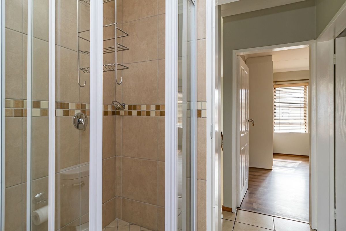 A bathroom featuring a tiled shower enclosure with a mosaic border, leading into a hallway with wood-look flooring.