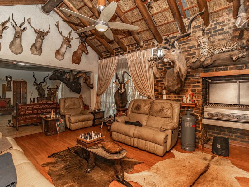 Spacious living area featuring a rustic thatched roof with exposed timber beams, brick fireplace, built-in braai, and laminate flooring.