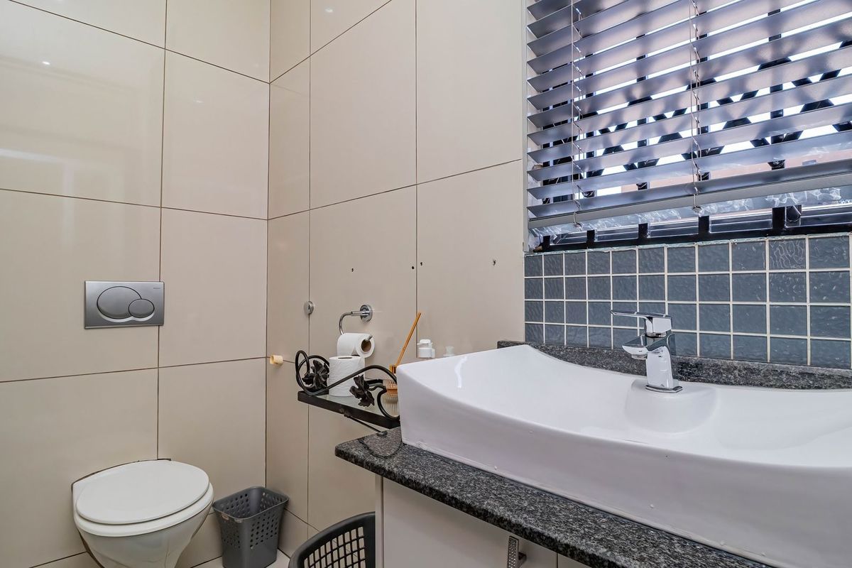 A compact bathroom featuring a modern vessel sink on a granite vanity, wall-mounted toilet, and neutral wall tiling with a window fitted with venetian blinds.