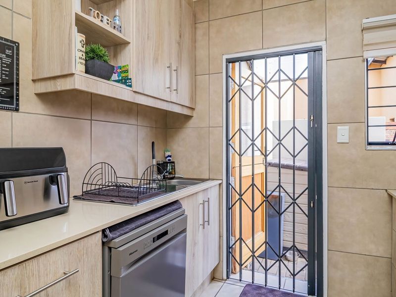 A functional scullery featuring light wood-finish cabinetry, tiled walls, a stainless steel sink, and a secure exterior door with a metal security gate.