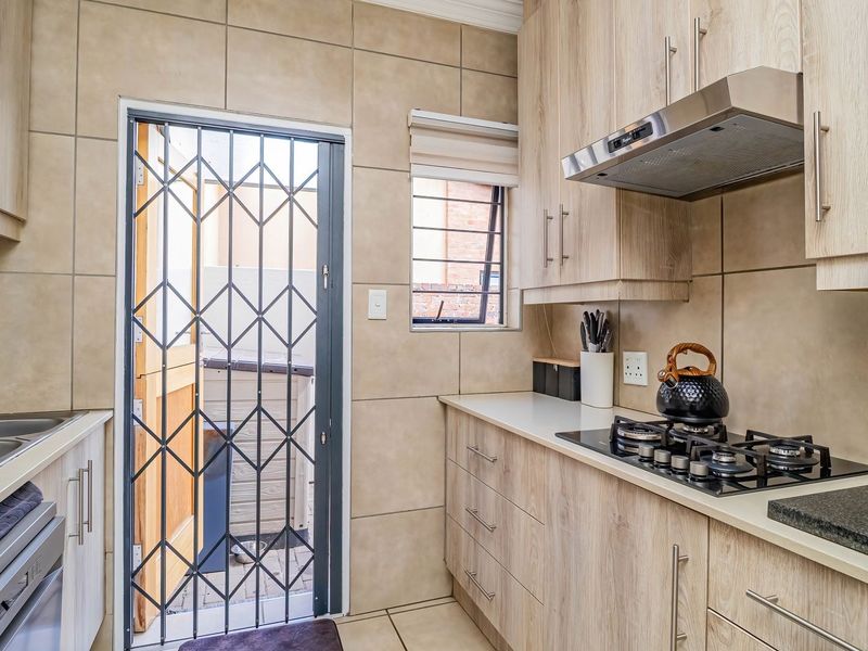 A well-maintained kitchen featuring light wood-finish cabinetry, stone countertops, a gas hob with extractor fan, and direct access to an exterior area via security gate.