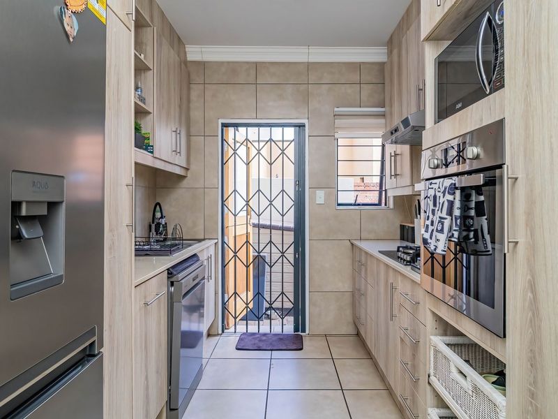 A well-maintained galley-style kitchen featuring light wood-finish cabinetry, tiled flooring, built-in oven and microwave, and a secure rear exit door.