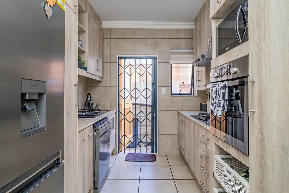 A well-maintained galley-style kitchen featuring light wood-finish cabinetry, tiled flooring, built-in oven and microwave, and a secure rear exit door.