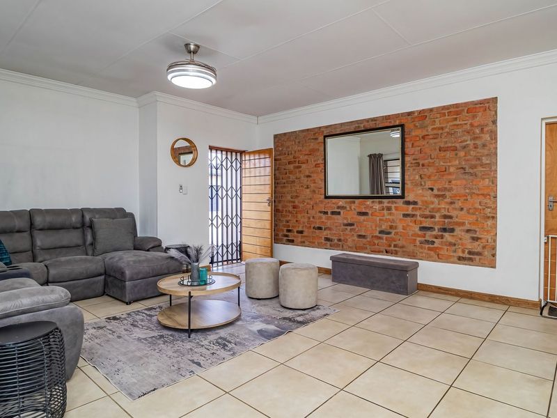Spacious living room featuring a prominent exposed brick accent wall, neutral ceramic floor tiles, and a ceiling fan, providing a comfortable and well-maintained living space.