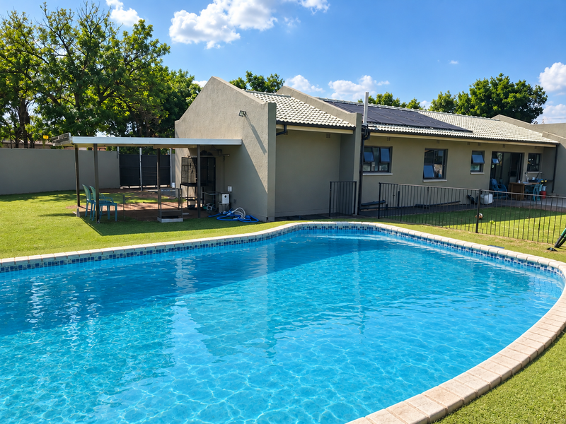 A well-maintained backyard featuring a clear swimming pool with solar heating panels, a covered patio area, and secure perimeter fencing.
