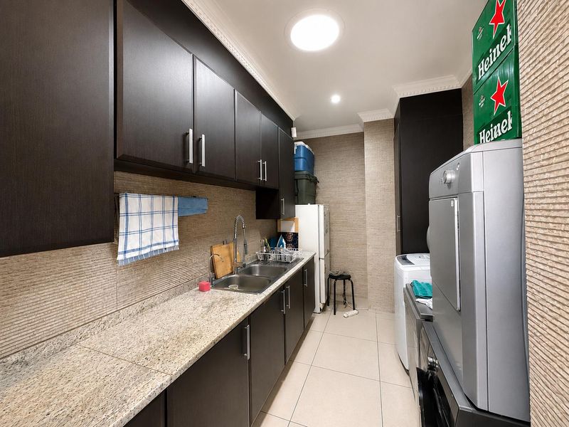A functional scullery featuring dark cabinetry, granite countertops, a double stainless steel sink, and tiled finishes, providing ample storage and workspace.