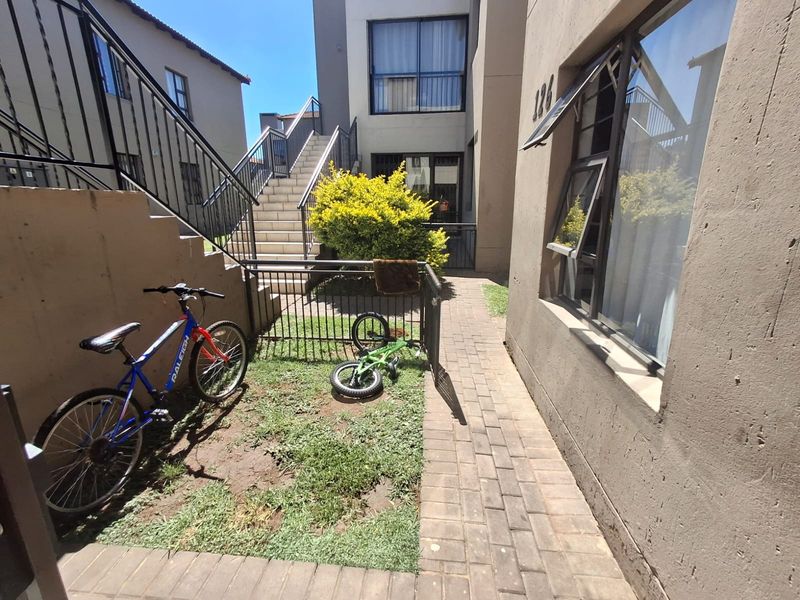 A rear exterior view of an apartment complex featuring a paved walkway, external staircase, and small garden area with metal security fencing.