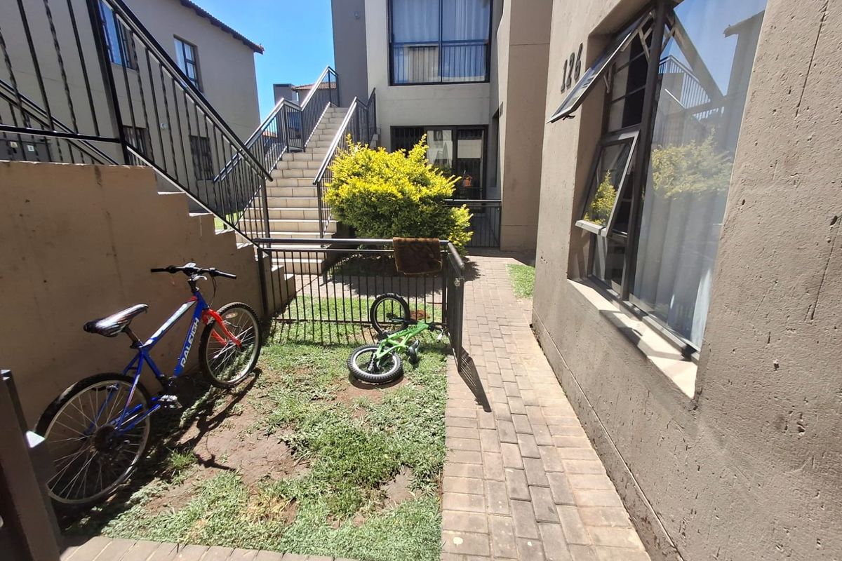 A rear exterior view of an apartment complex featuring a paved walkway, external staircase, and small garden area with metal security fencing.
