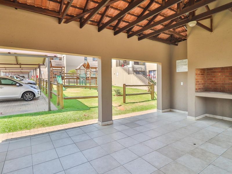 A covered outdoor patio area featuring tiled flooring, exposed timber roof trusses, and a built-in brick braai, overlooking the complex garden and parking area.