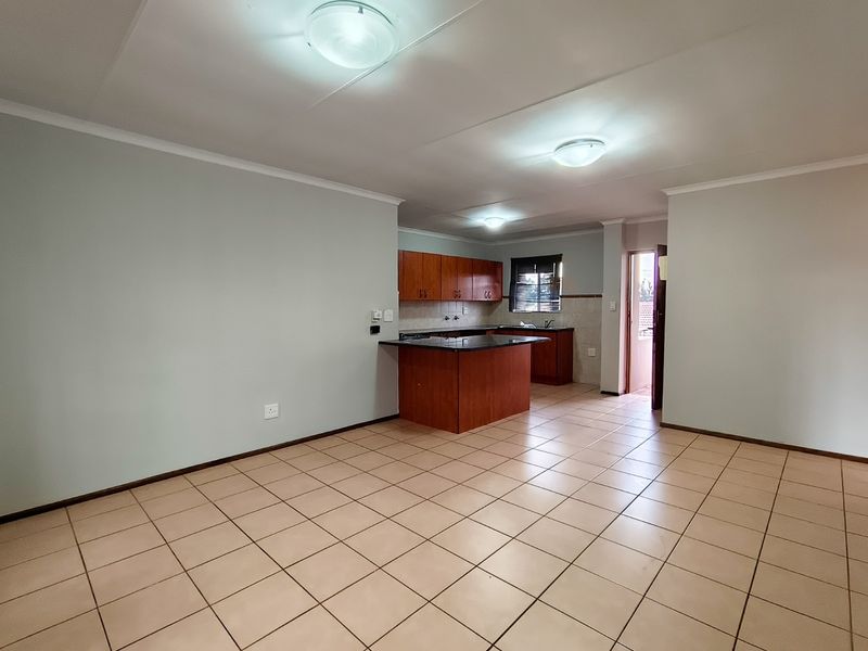 An open-plan living and kitchen area featuring tiled flooring, wooden cabinetry, granite countertops, and a breakfast bar in a clean, vacant space.