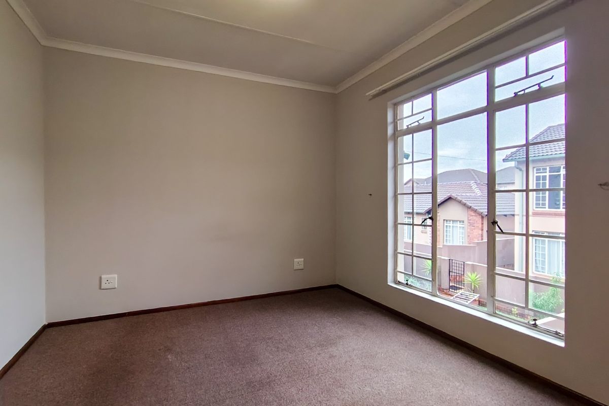 A bright, empty bedroom featuring neutral walls, carpeted flooring, and a large multi-pane window providing natural light and a view of the surrounding neighborhood.