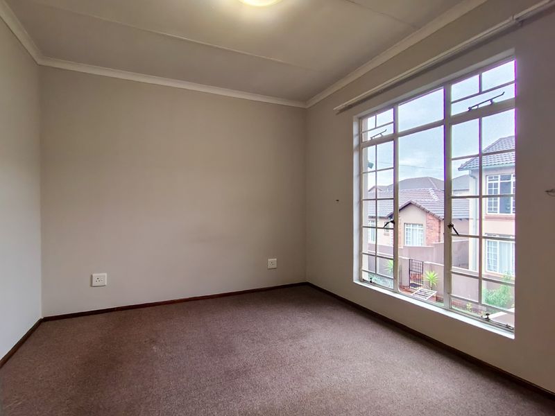 A bright, empty bedroom featuring neutral walls, carpeted flooring, and a large multi-pane window providing natural light and a view of the surrounding neighborhood.
