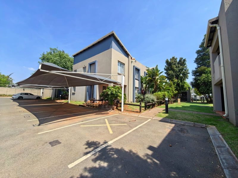 Exterior view of a townhouse complex featuring a paved driveway, designated parking bays with shade cloth carports, and established garden areas.