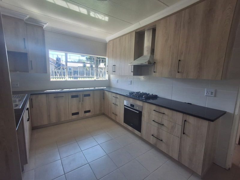 A modern, clean kitchen featuring light wood-grain cabinetry, dark stone countertops, built-in oven, gas hob, and an extractor fan with neutral tiled flooring.