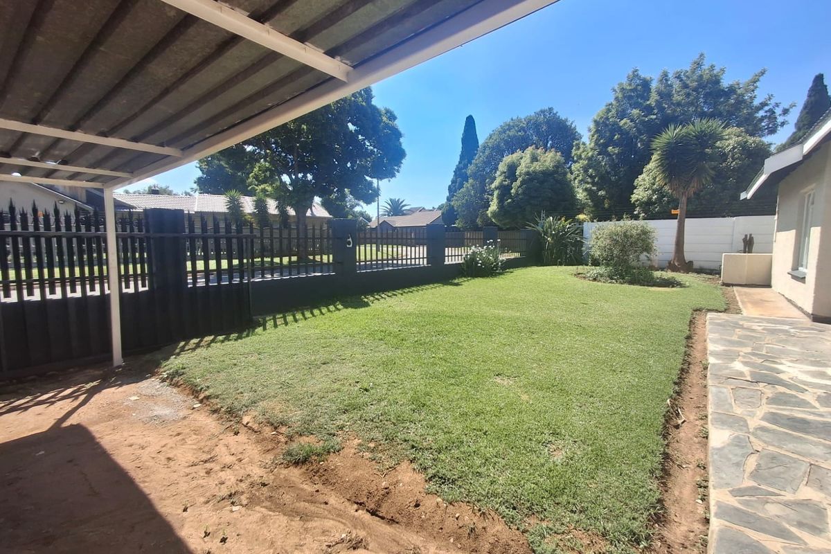 A spacious rear garden featuring a manicured lawn, paved walkway, and a covered carport area enclosed by steel perimeter fencing.