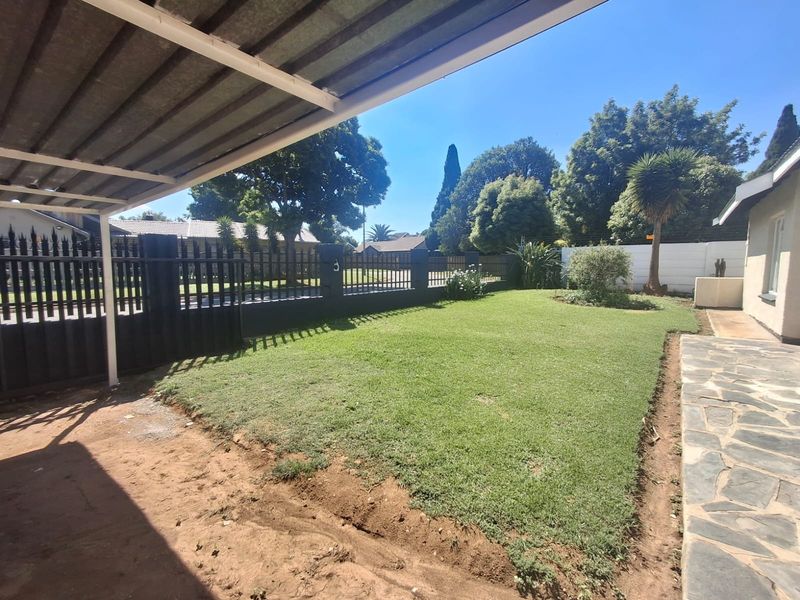 A spacious rear garden featuring a manicured lawn, paved walkway, and a covered carport area enclosed by steel perimeter fencing.