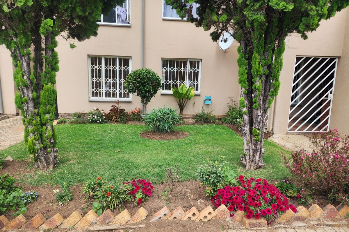A well-maintained rear garden area featuring established trees, flower beds with brick edging, and security features on the apartment windows and entrance.