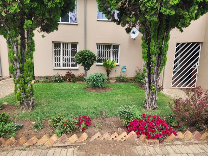 A well-maintained rear garden area featuring established trees, flower beds with brick edging, and security features on the apartment windows and entrance.