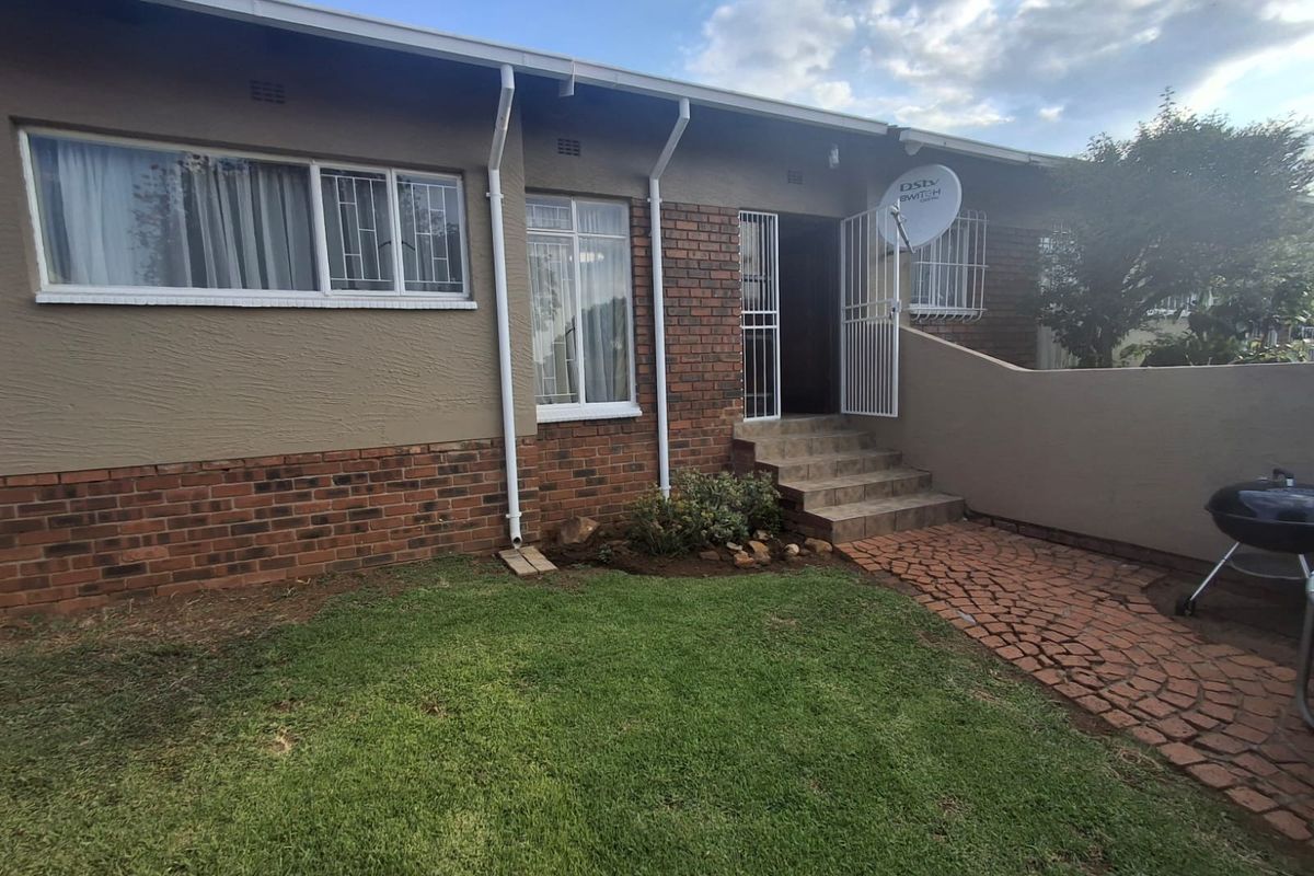 Rear exterior view of the townhouse featuring a brick and plaster facade, paved walkway, small lawn, and secure entrance with security gate.