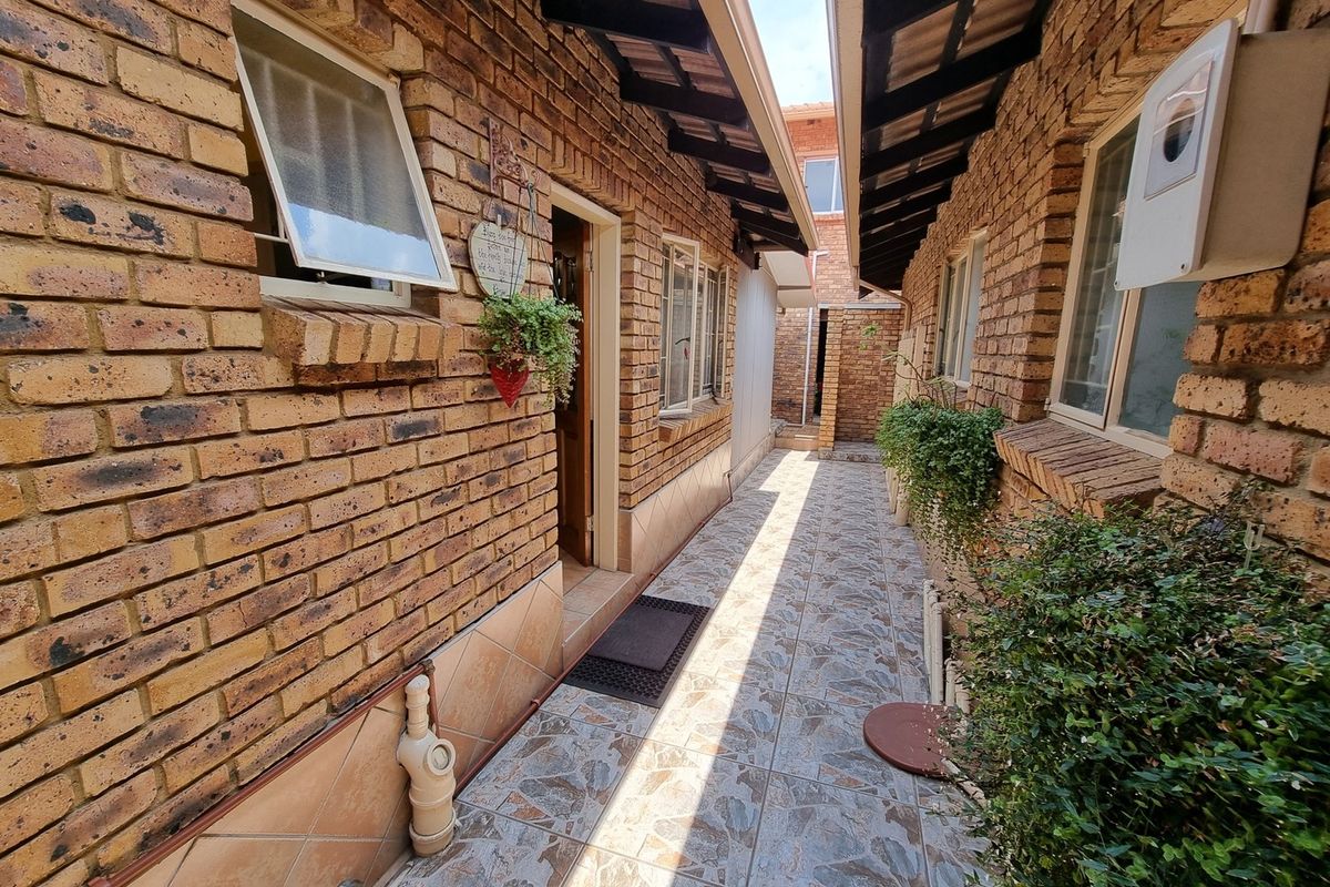 A narrow exterior walkway between brick building sections, featuring tiled flooring, exposed brick walls, and a covered roof structure.