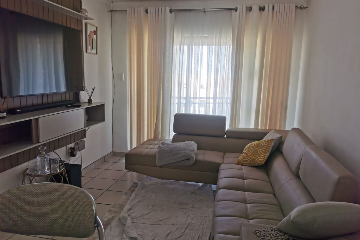 A cozy living room featuring tiled flooring and a built-in media unit, with large windows dressed in curtains allowing natural light into the space.
