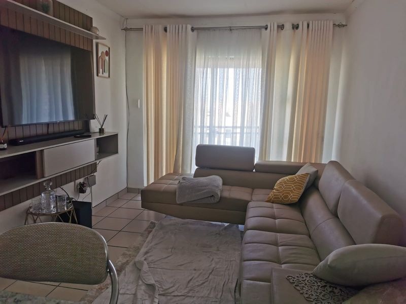 A cozy living room featuring tiled flooring and a built-in media unit, with large windows dressed in curtains allowing natural light into the space.