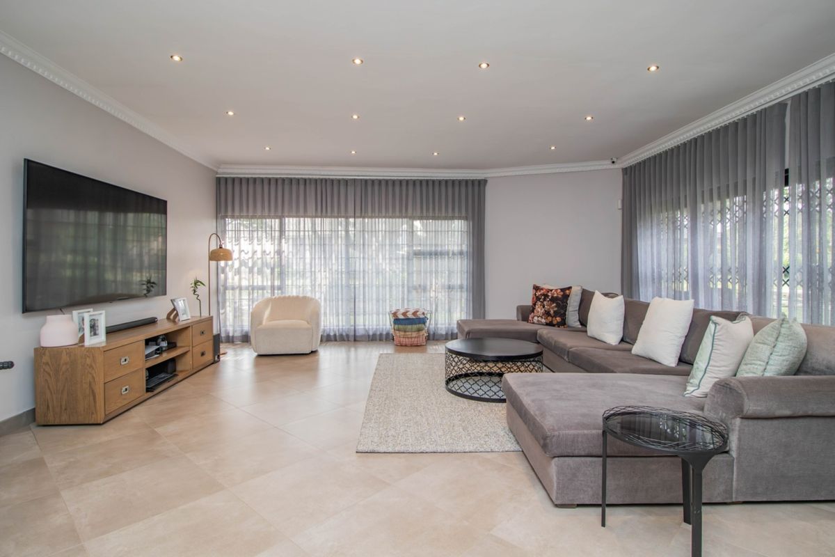 A spacious, well-lit living room featuring neutral tiled flooring, recessed ceiling lighting, crown molding, and large windows with floor-to-ceiling curtains.
