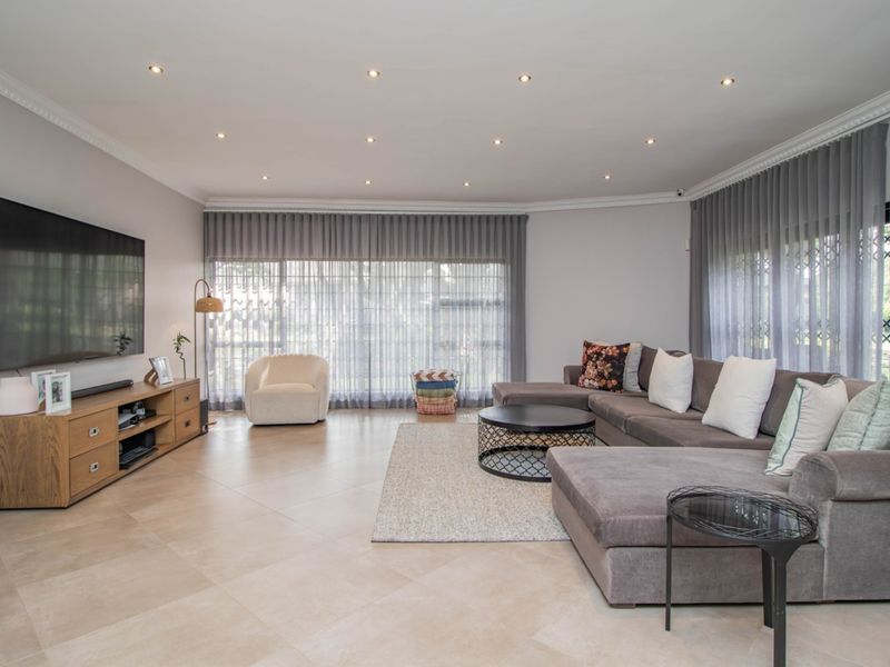 A spacious, well-lit living room featuring neutral tiled flooring, recessed ceiling lighting, crown molding, and large windows with floor-to-ceiling curtains.