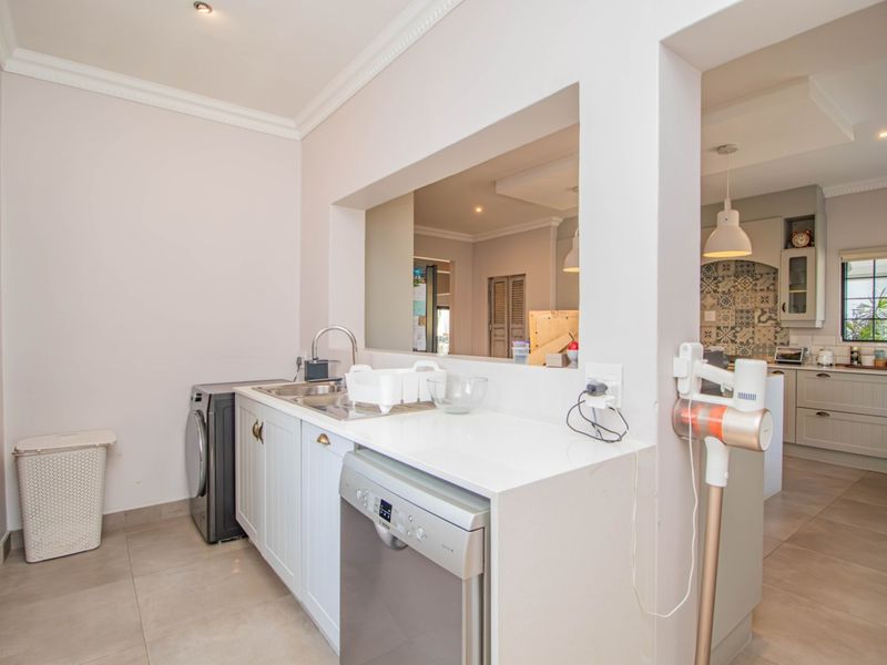 A functional scullery area featuring built-in cabinetry, stone countertops, a stainless steel sink, and tiled flooring, with a convenient pass-through window to the kitchen.
