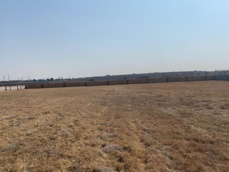 A large, vacant residential plot of land featuring dry grass and a perimeter brick boundary wall.