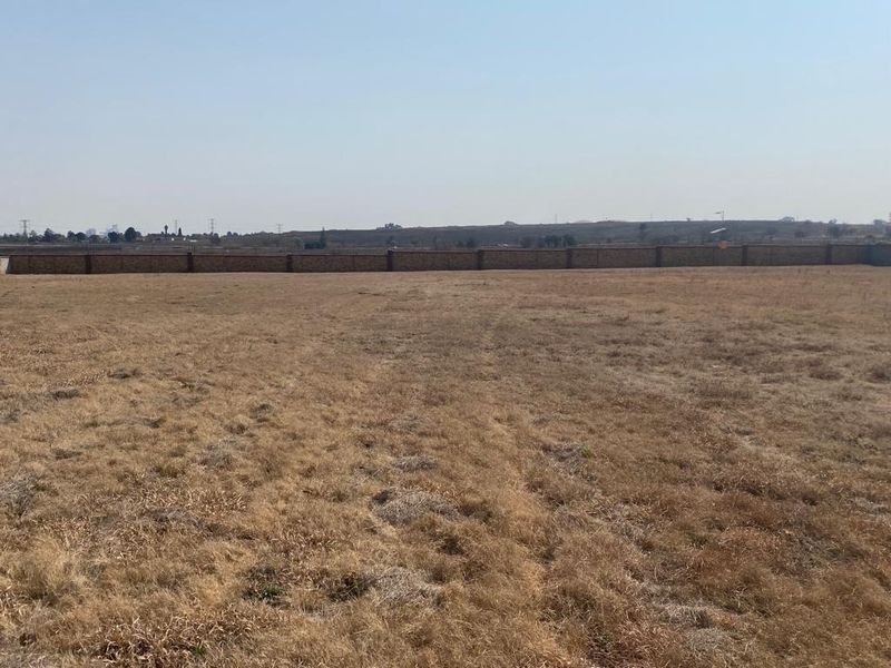A wide, vacant residential plot featuring dry grass and a perimeter brick boundary wall.
