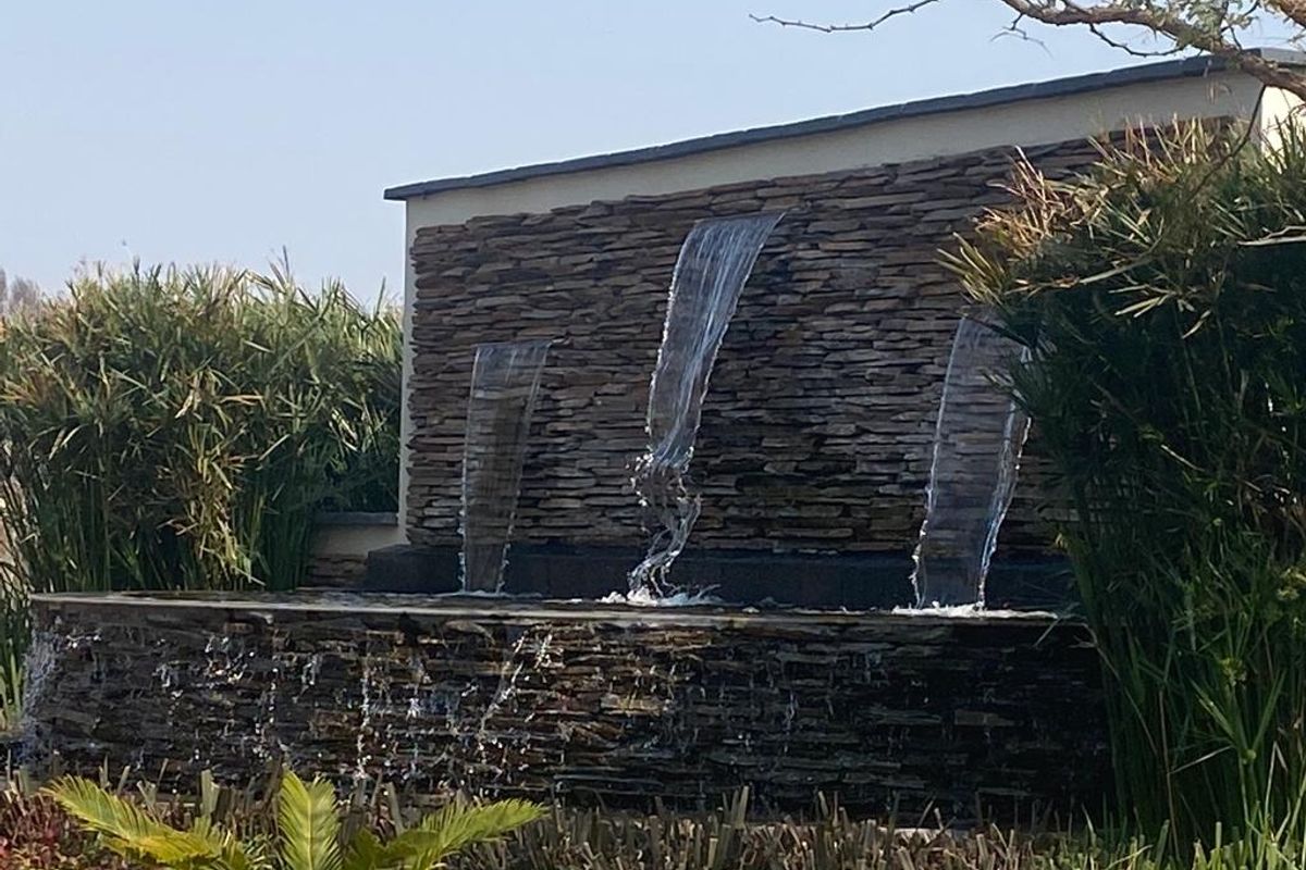 A beautifully maintained stone-clad water feature with three cascading waterfalls, set within a lush, landscaped garden environment.