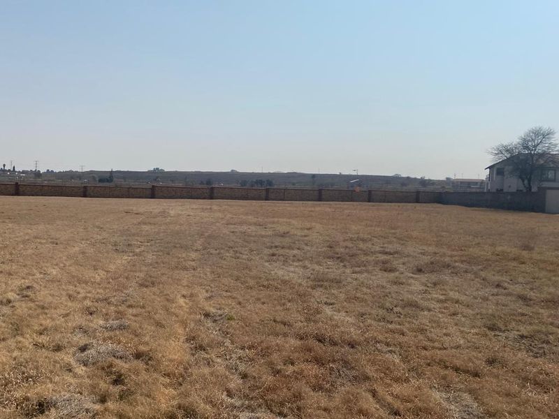 A vacant residential plot featuring dry grass and a perimeter boundary wall, offering a clear view of the surrounding landscape.