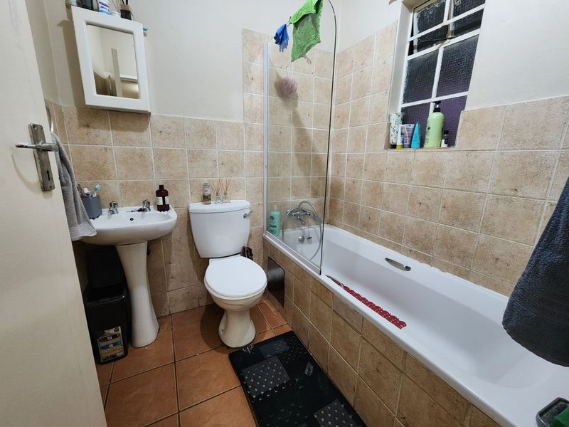 A functional bathroom featuring a pedestal basin, toilet, and a bathtub with a glass shower screen, finished with neutral wall and floor tiles.