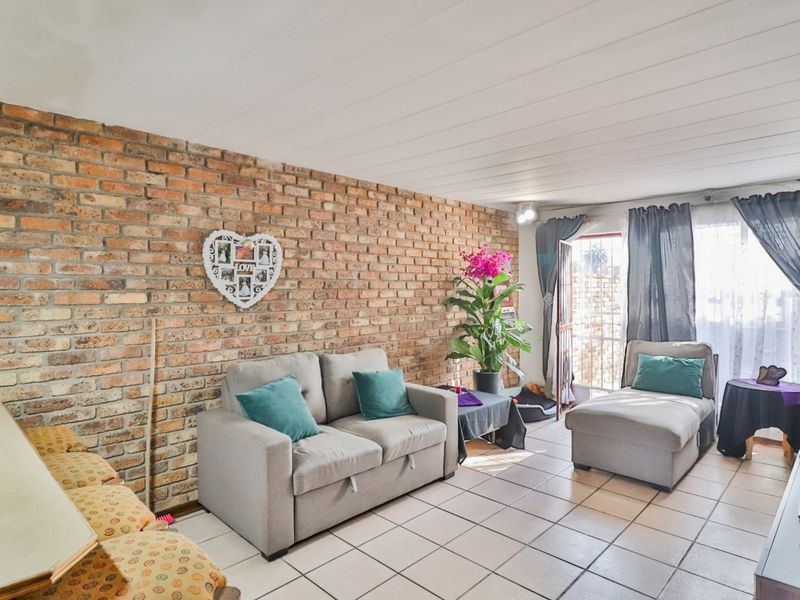 A living room featuring an exposed brick feature wall, tiled flooring, and a panelled ceiling with direct access to an outdoor area.