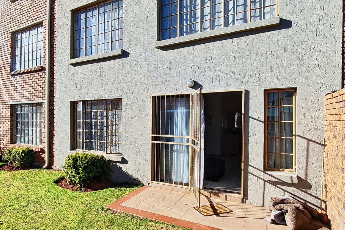 Rear view of an apartment building featuring a plastered facade, brick accents, tiled patio area, and security gates on windows and doors.