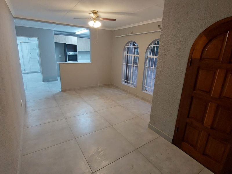 An open-plan living area featuring tiled floors, arched windows with security bars, and a view into the adjacent kitchen with built-in cabinetry.