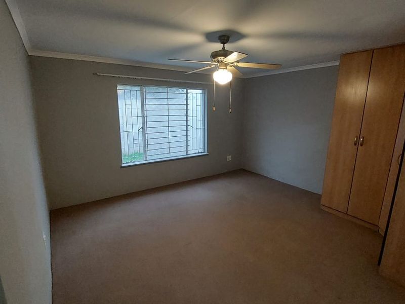A spacious, empty bedroom featuring neutral walls, carpeted flooring, built-in wooden cupboards, and a ceiling fan with security bars on the window.