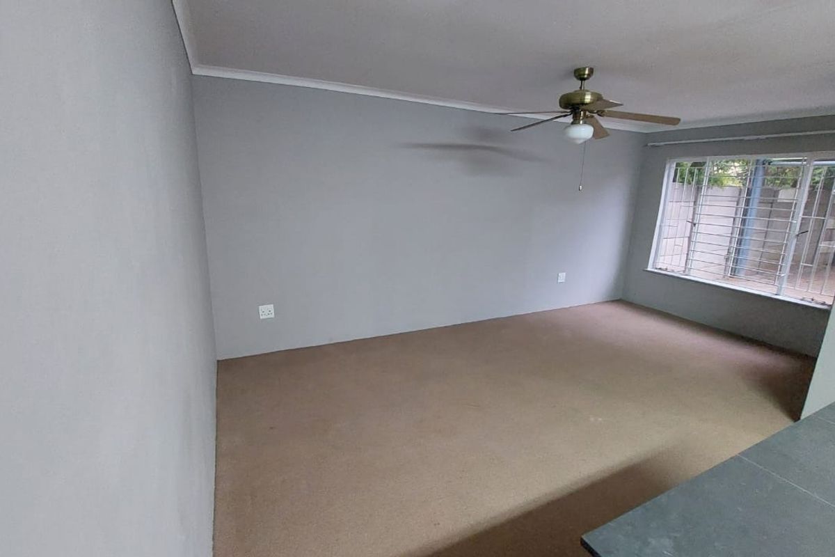 An empty living room featuring neutral grey walls, carpeted flooring, and a ceiling fan, with security bars installed on the window.