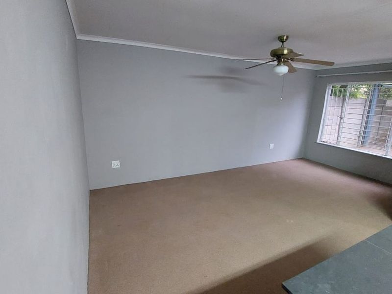 An empty living room featuring neutral grey walls, carpeted flooring, and a ceiling fan, with security bars installed on the window.