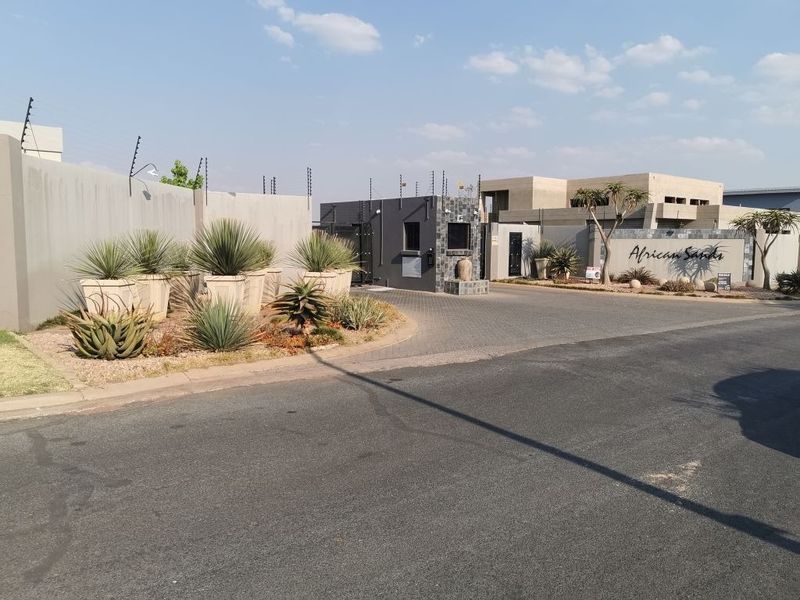 The entrance to African Sands estate featuring a modern security gatehouse, paved driveway, and perimeter boundary walls with electric fencing.