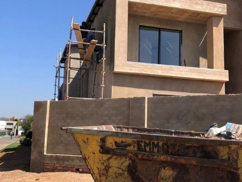 Front elevation of a house under construction, featuring a plastered facade, balcony with sliding doors, and a perimeter wall.