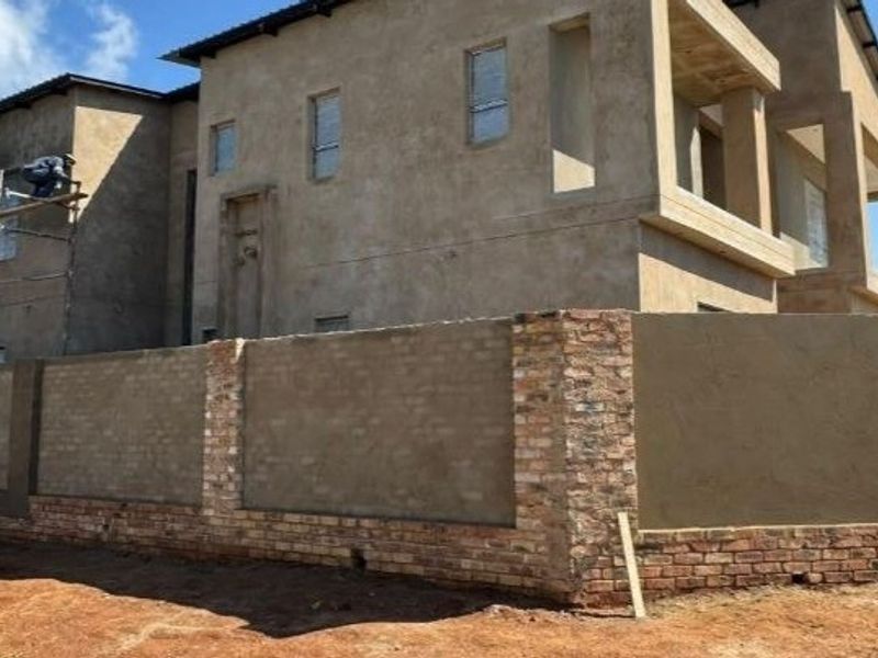 Exterior view of a double-story house under construction, featuring a brick boundary wall and ongoing plastering work on the facade.