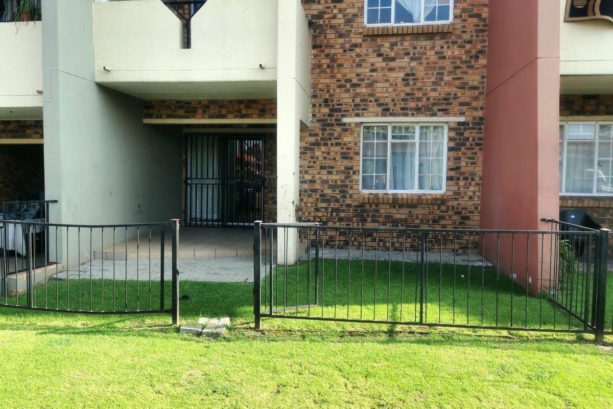 Ground floor apartment exterior featuring a private paved patio, small lawn area, and secure metal fencing, with a balcony above.