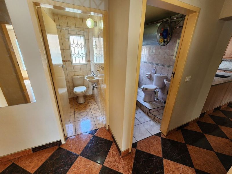A hallway providing access to two separate bathrooms, featuring tiled flooring and functional bathroom fixtures with an exposed water heater.