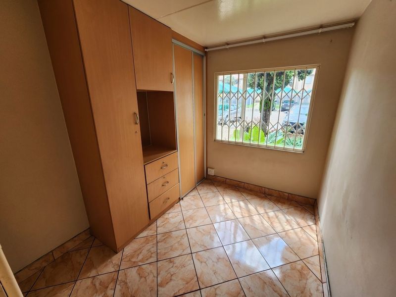 A compact bedroom featuring built-in wooden cabinetry, ceramic tiled flooring, and a window fitted with security bars.