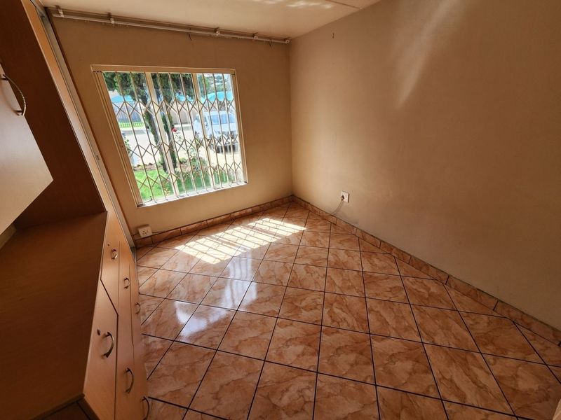 A bright, empty bedroom featuring tiled flooring, built-in cupboards, and a window fitted with a security gate.