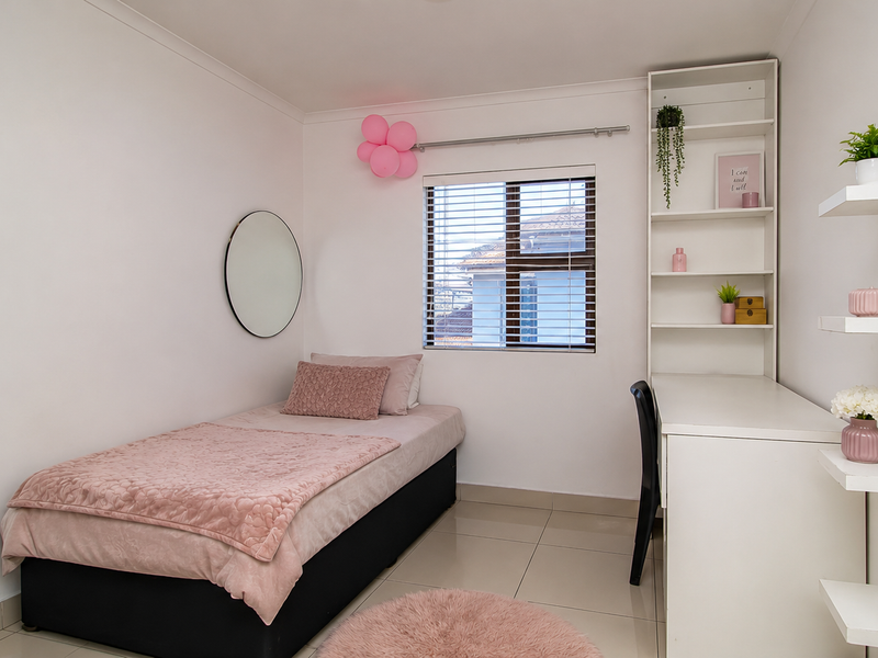 A well-maintained bedroom featuring neutral tiled flooring, a built-in desk with integrated shelving, and a window fitted with venetian blinds.