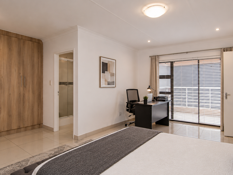 Spacious bedroom featuring built-in wooden cupboards, tiled flooring, and a sliding door leading to a balcony, with an adjoining en-suite bathroom.